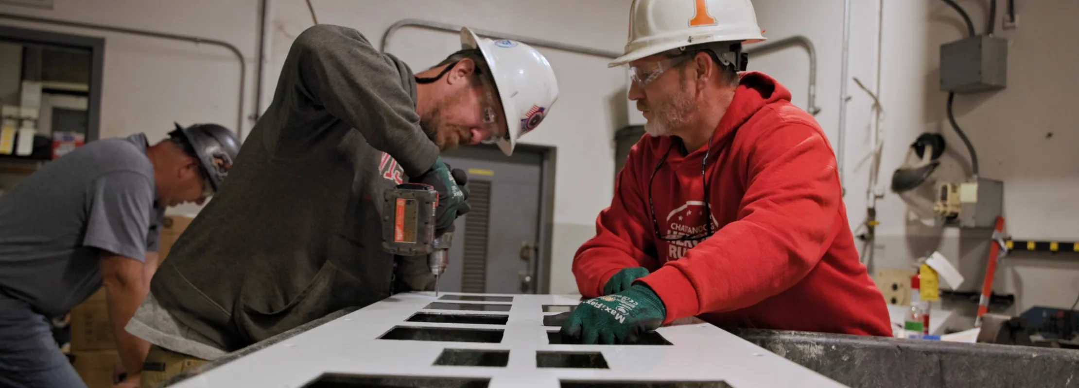 Two construction workers wearing hard hats working on a white panel with multiple square cutouts indoors.