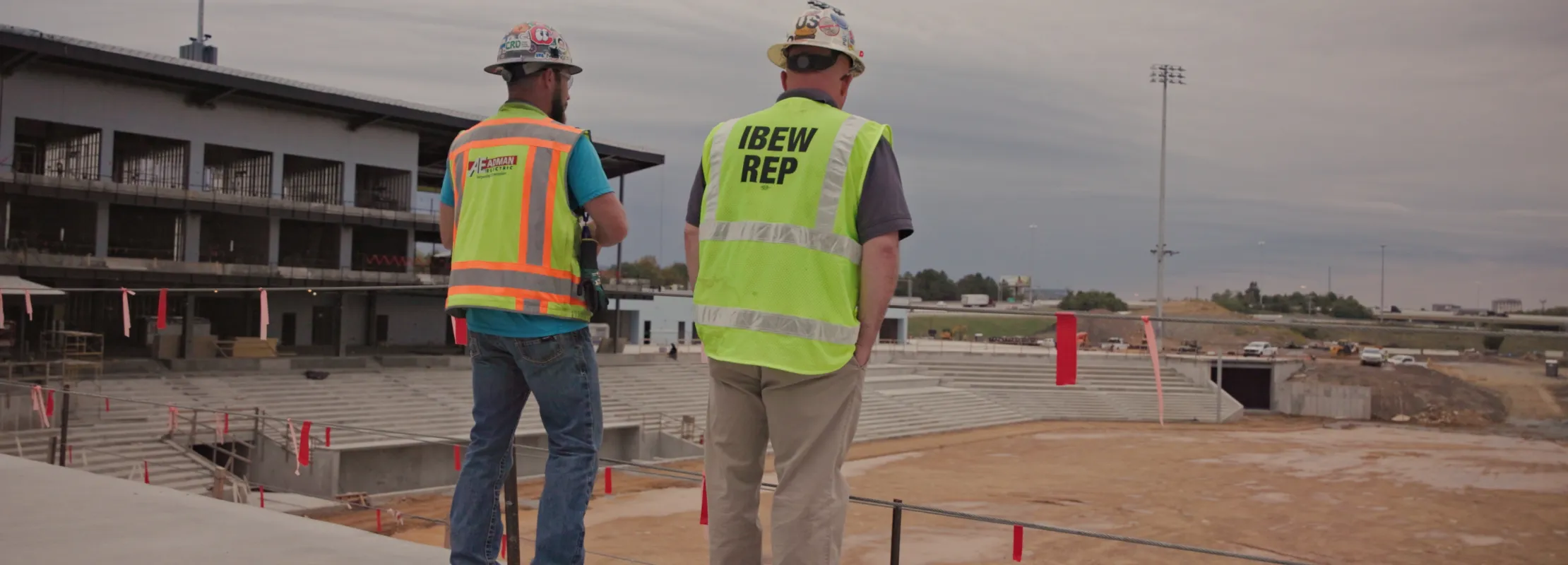 Two construction workers in safety vests and helmets inspecting a large stadium construction site outdoors.