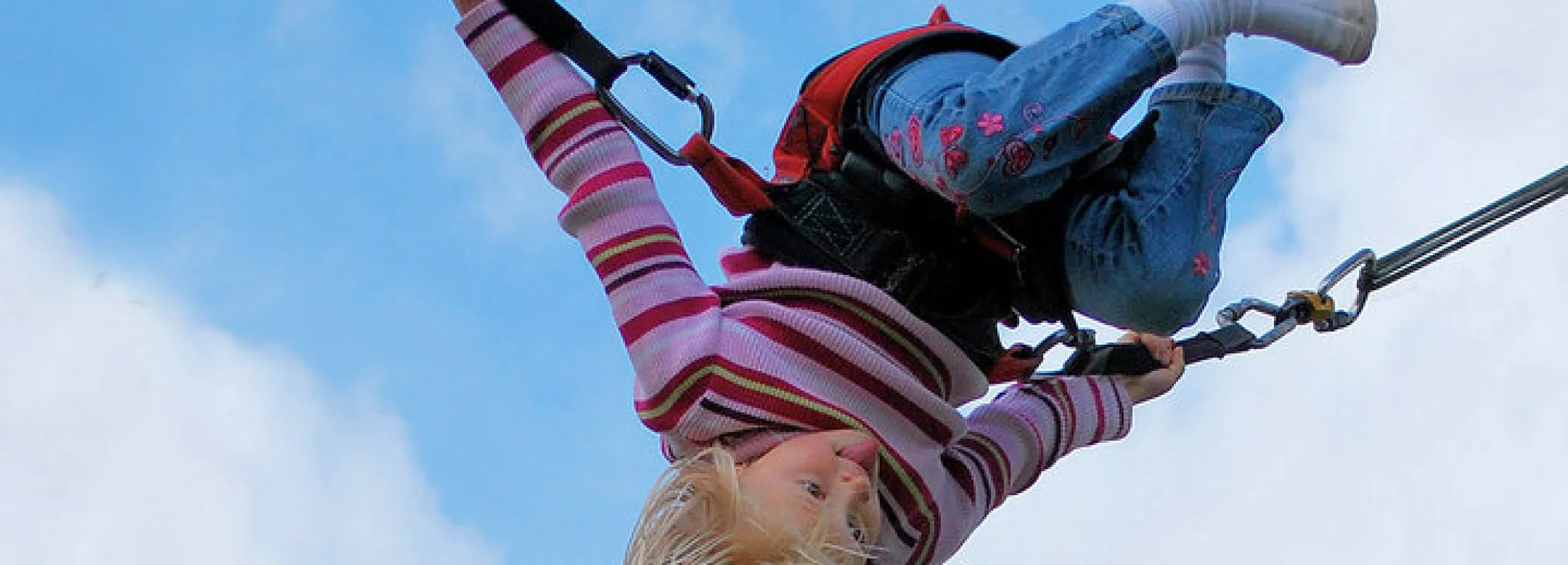 Young child with blonde hair swinging upside down on a harness against a blue sky.
