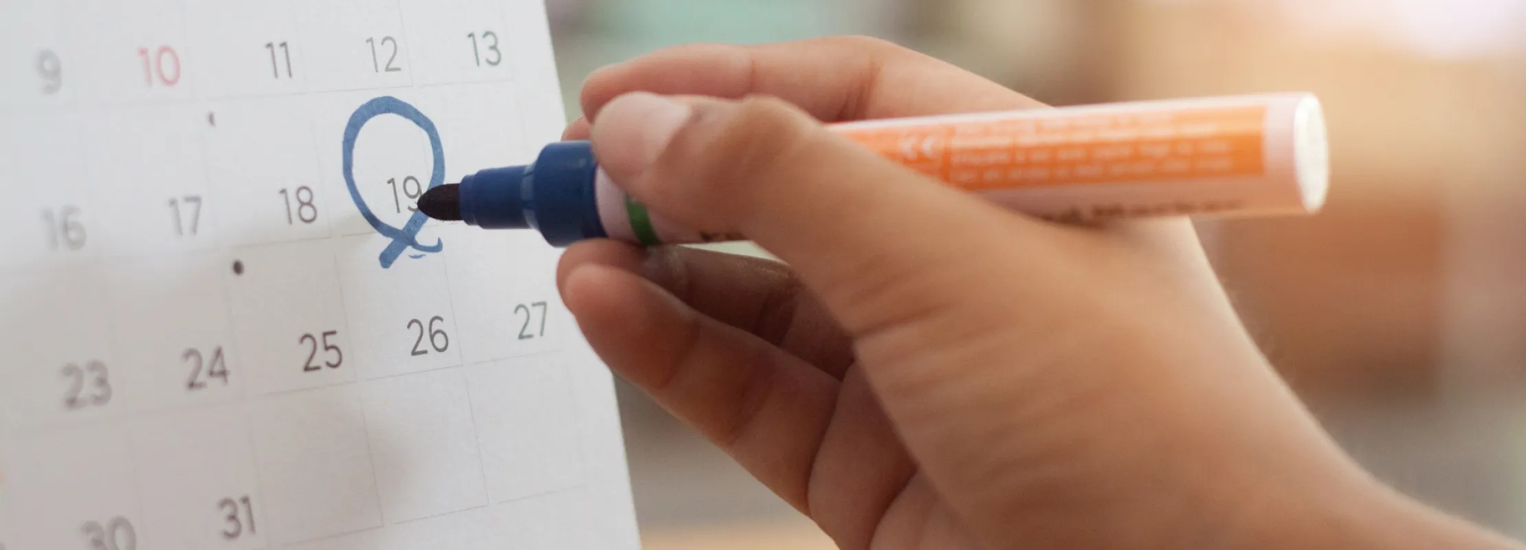 Hand marking a circle around the date 19 on a desk calendar with a blue marker.