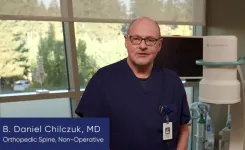 Doctor B. Daniel Chilczuk in navy scrubs standing in a medical office with equipment and trees outside the window.