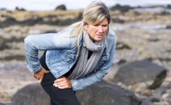 Mature woman outdoors holding hip in pain on rocky shore, wearing denim jacket and gray scarf.