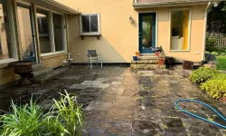 Thumbnail control image for Sunny backyard patio with stone tiles, scattered plants, garden hose, and yellow house exterior with windows and doors.