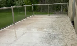 Thumbnail control image for Wet concrete patio with glass railing overlooking lush green trees and a blue hose on the floor.