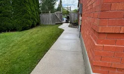 Thumbnail control image for Concrete pathway alongside a red brick house with green lawn, tall evergreen trees, and a wooden gate under a cloudy sky.