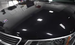 Thumbnail control image for Close-up of a shiny black car hood with a Saab emblem, grille, and headlight under bright indoor lights
