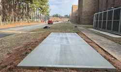 Thumbnail control image for Newly poured concrete sidewalk section beside a brick building under a cloudy sky with trees lining the road.
