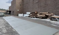 Thumbnail control image for Newly poured concrete sidewalk next to a brick warehouse with stacked cardboard and pallets outside.