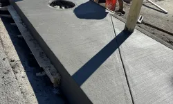 Thumbnail control image for Construction workers installing freshly poured concrete sidewalk curb with circular hole and yellow bollards.