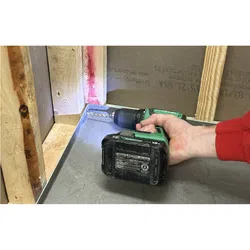 Hand holding green cordless drill with LED light, drilling into metal surface near wooden studs in construction.