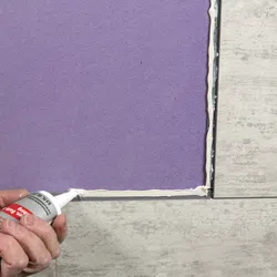 Hand applying white adhesive or sealant along the edge of a purple tile on a tiled floor corner.