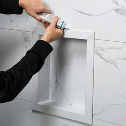 Person applying sealant to corner of recessed marble tiled shower niche on wall installation.