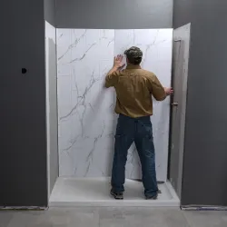 Man installing white marble-patterned wall panels in a shower with gray walls and a white base