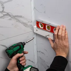 Person installing wall shelf on marble tiled wall using electric screwdriver and spirit level for alignment.