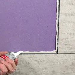 Hand applying white adhesive sealant around edge of purple tile on beige floor during installation