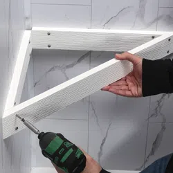 Person installing a white wooden shelf frame with a power drill on a marble wall background.