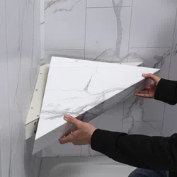 Person installing a white marble-patterned triangular wall shelf in a marble-tiled corner space.