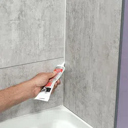 Hand applying white waterproof sealant caulk to bathtub corner between tiled walls.