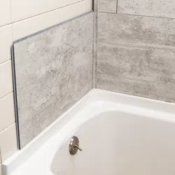 Close-up of bathtub corner with beige ceramic and gray stone-patterned wall tiles and a silver drain stopper.