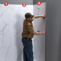 Man installing a glass shower door in a modern bathroom with white marble tiled walls and gray ceiling.