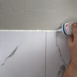 Hand applying white caulk along the edge where marble tiles meet a painted wall for sealing.
