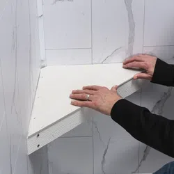 Person installing a white triangular wall shelf against a marble tiled wall indoor.