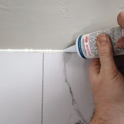 Hand applying white caulk between marble tiles and wall for sealing and finishing touch in home improvement.