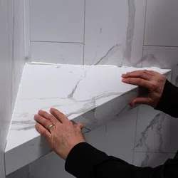 Hands installing a white marble corner shelf with gray veining on a tiled wall.