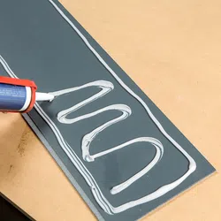 Hand applying white silicone sealant in a zigzag pattern on a dark grey sheet on a wooden surface.