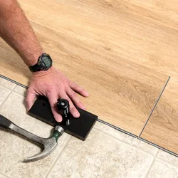 Close-up of hands installing wooden laminate flooring over tiled floor using hammer and tapping block.