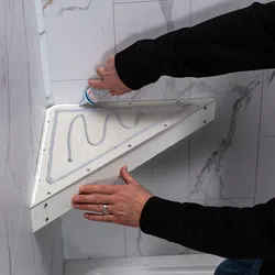 Person applying adhesive to a triangular wooden shelf before mounting it on a tiled wall corner in a bathroom.
