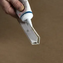 Hand applying white adhesive sealant from a tube onto a white plastic corner trim piece against a brown background