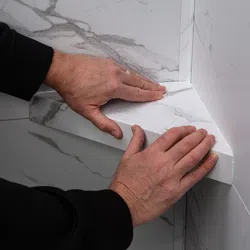 Hands installing a white marble-patterned shelf or ledge in a corner against matching wall tiles.