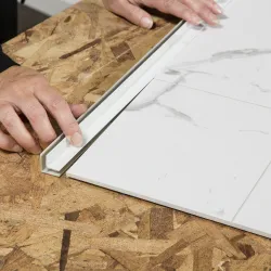 Hands installing a white marble-pattern tile with a white trim piece on a plywood floor surface.