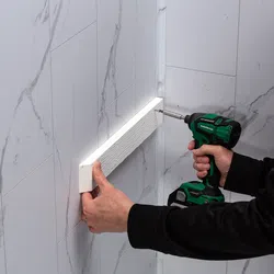 Person using a green cordless drill to install a white textured wall trim on marble tile wall corner.