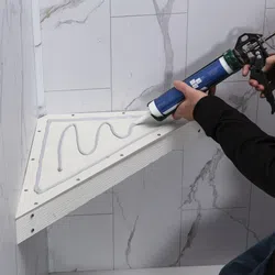 Person applying adhesive with caulking gun to attach a triangular wooden shelf in a bathroom corner