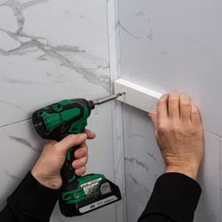 Hand installing white trim on marble tile wall corner using green cordless drill screwdriver.