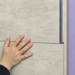 Hand installing large gray wall tile panels against a purple background, showing smooth texture and precise fitting.