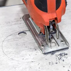 Close-up of an electric jigsaw cutting a circular hole in a wooden board with pencil markings.