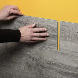 Hands installing gray wood-look vinyl flooring panels on a yellow surface with a tongue and groove fit.