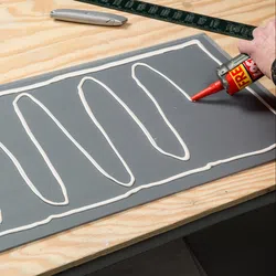 Applying white construction adhesive in zigzag pattern on gray panel on wooden workbench with tools nearby