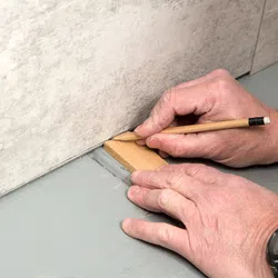 Hands marking a wooden tile with a pencil on a gray floor near a tiled wall during installation.