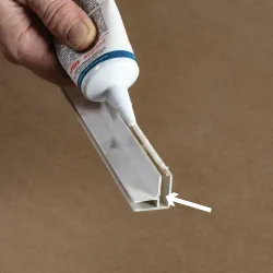 Hand applying white adhesive glue to a plastic joint piece for repair or assembly on a brown background