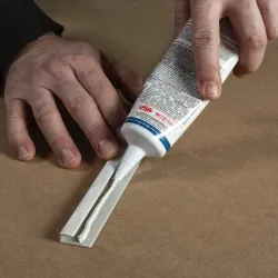 Person applying white adhesive from a tube onto a hinge on a brown surface.