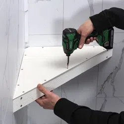 Person using a green cordless drill to fasten a white triangular shelf against a marble wall.