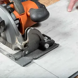 Close-up of a circular saw cutting a tile with an orange handle and black grip on a work surface.