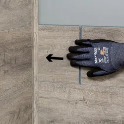 Hand wearing a protective glove pointing at a wooden tile with a black arrow on a tiled surface.