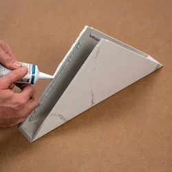 Hands applying adhesive sealant to the edge of a white marble-patterned triangular shelf on a brown surface.