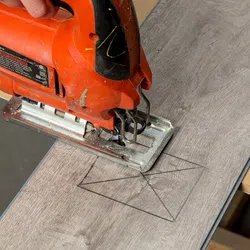 Close-up of a hand using an orange jigsaw to cut a marked pattern on gray laminate wood flooring.
