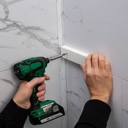 Person using a green cordless drill to screw a white wooden board into a marble wall corner.